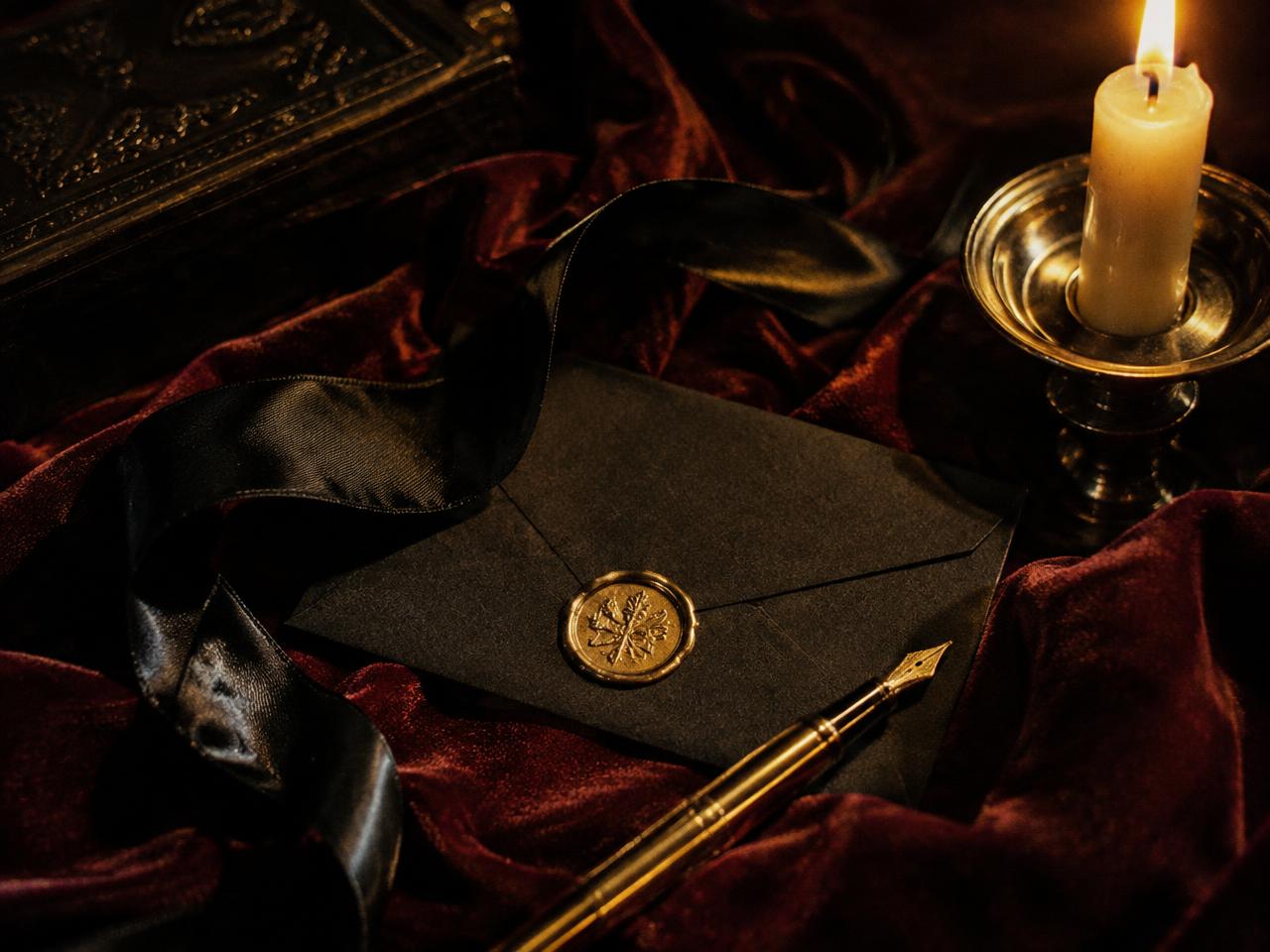 A black envelope sealed with gold wax beside a candle and fountain pen